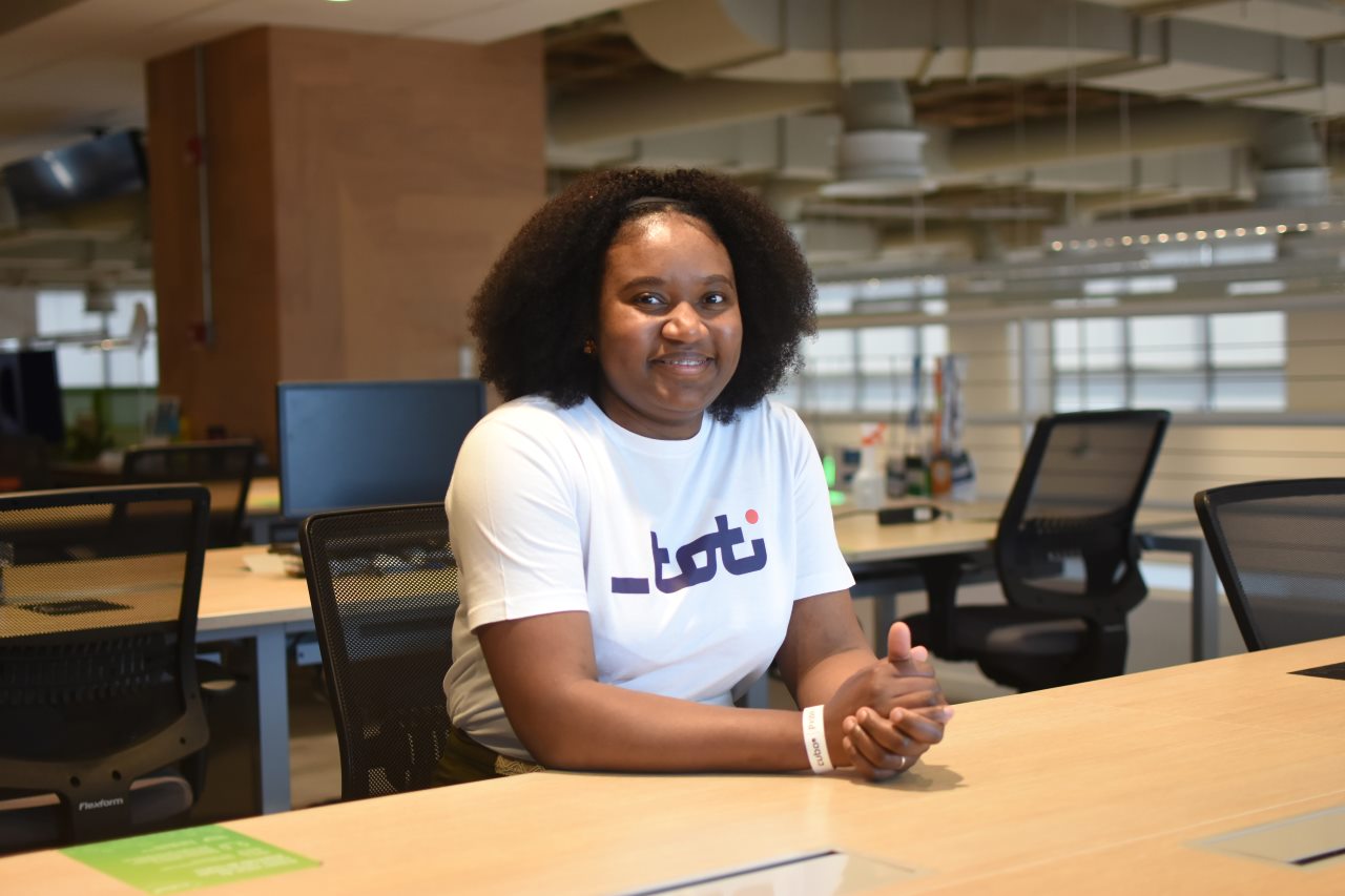 Foto de uma mulher sorridente sentada em uma mesa de escritório, vestindo uma camiseta branca com o logo da Toti. Ela está em um ambiente de trabalho moderno e iluminado.