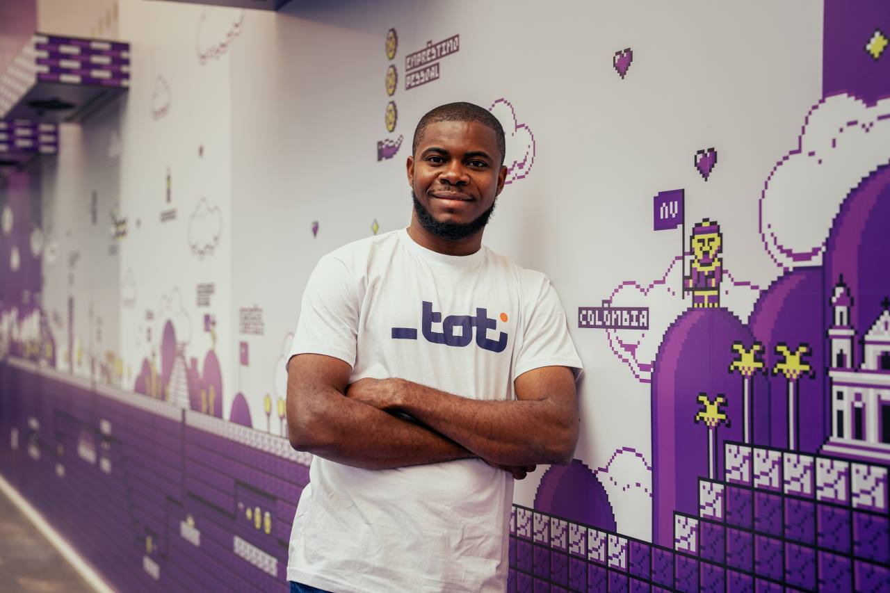 Foto de um homem sorrindo com os braços cruzados, vestindo uma camiseta branca com o logo da Toti, em frente a uma parede decorada com arte em pixel de temas urbanos e culturais.