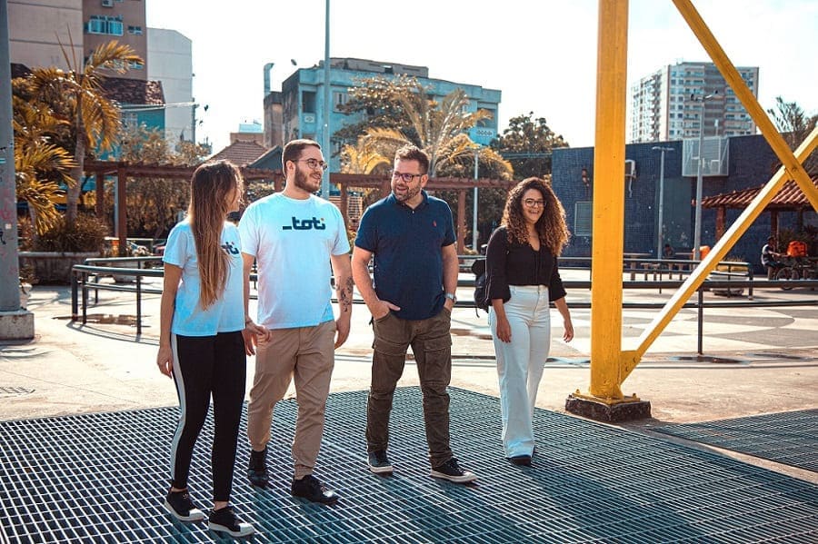 Quatro pessoas caminham em uma área aberta urbana. No grupo, estão dois homens e duas mulheres. Todos sorriem.