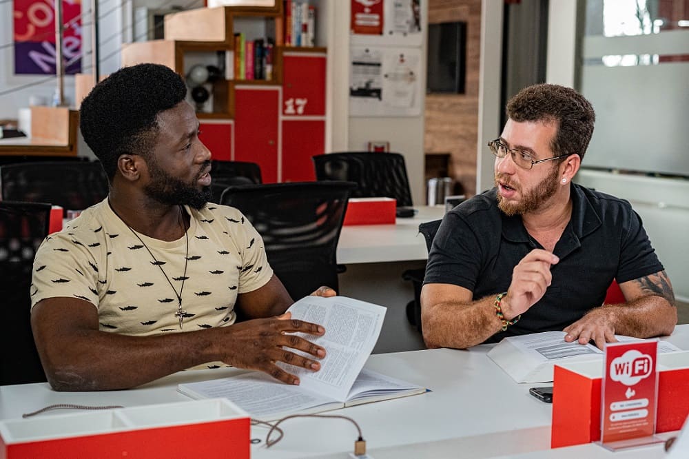 Dois homens conversam enquanto estão sentados em um ambiente de estudo e/ou trabalho. Os dois seguram livros em suas mãos.