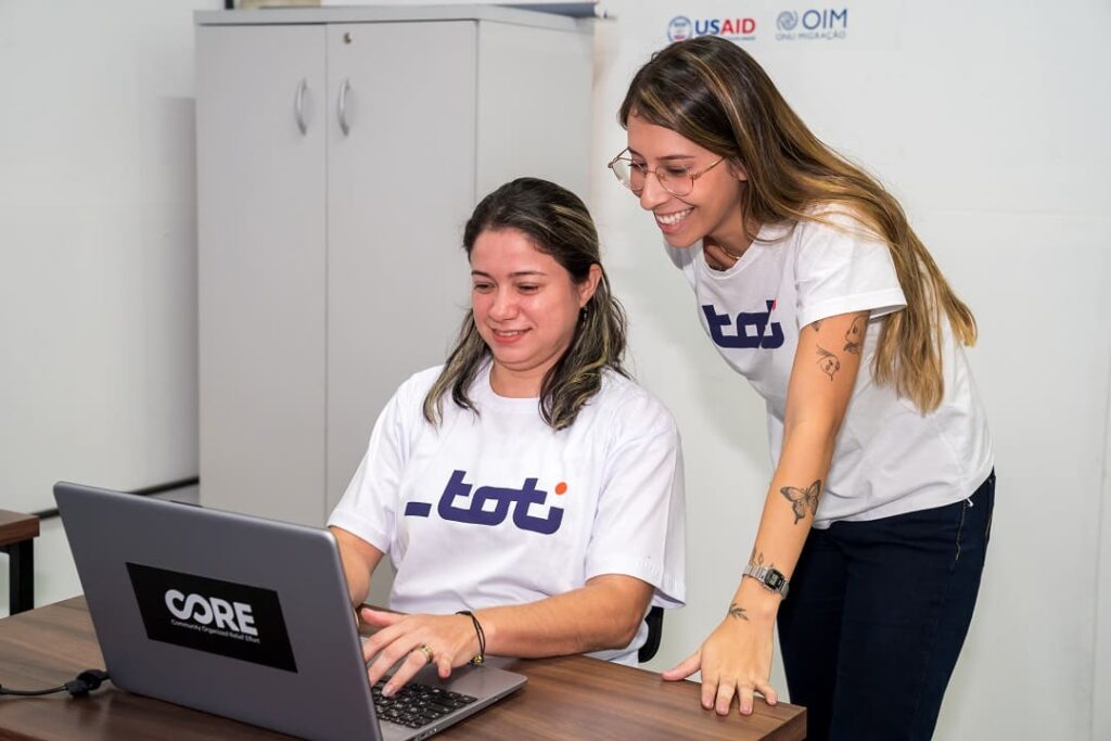 Uma mulher está sentada, trabalhando em um notebook. Ao seu lado, pé, está outra mulher. As duas sorriem e se mostram satisfeitas no ambiente de trabalho.