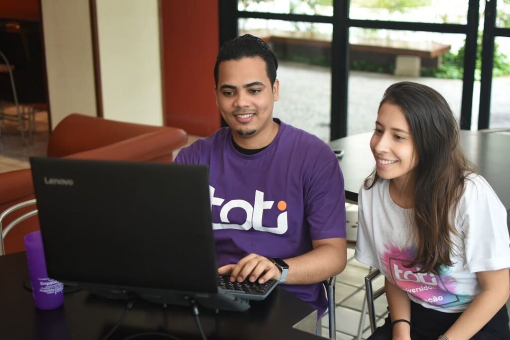 Um homem está sorrindo enquanto trabalha em seu notebook. Ao seu lado, está sentada uma mulher, também olhando para o notebook e sorrindo.