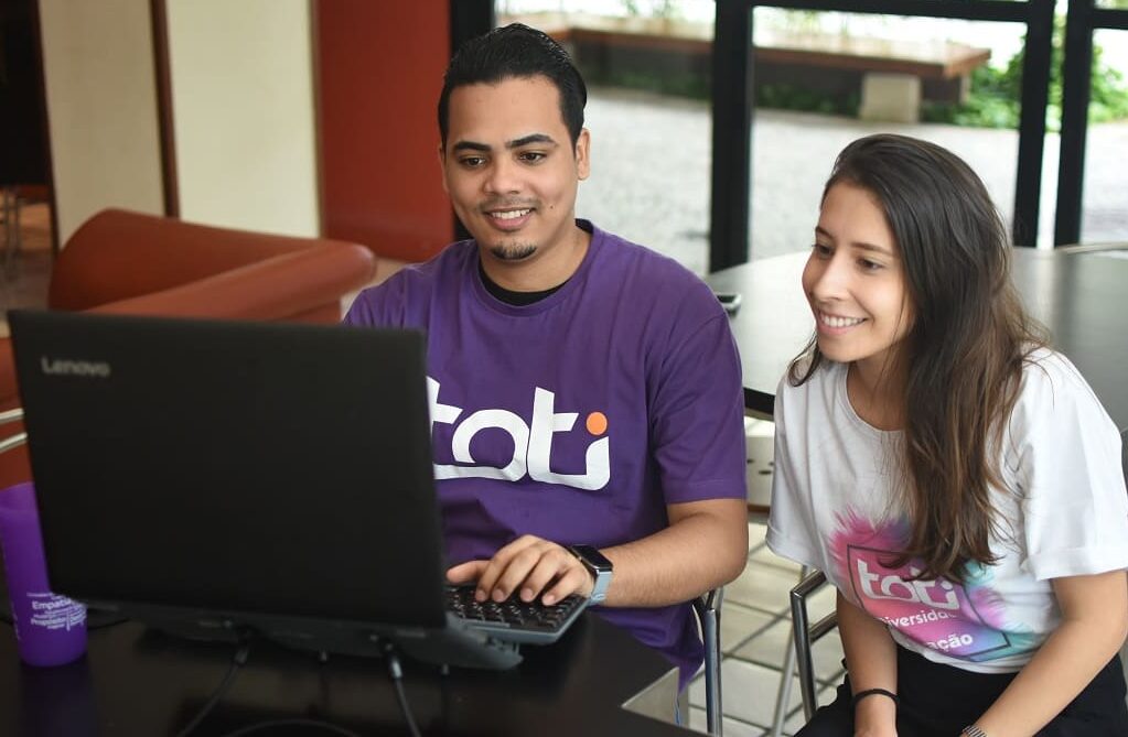 Um homem está sorrindo enquanto trabalha em seu notebook. Ao seu lado, está sentada uma mulher, também olhando para o notebook e sorrindo.