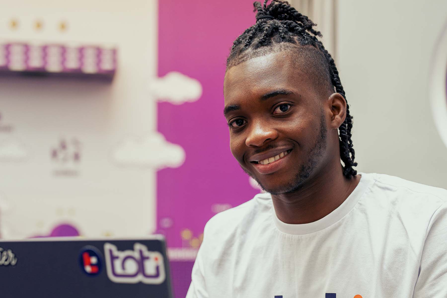 Foto de um jovem sorridente com cabelo trançado, vestindo uma camiseta branca. Ele está sentado, frente ao seu notebook, em um ambiente de escritório decorado com arte de pixel na parede ao fundo.