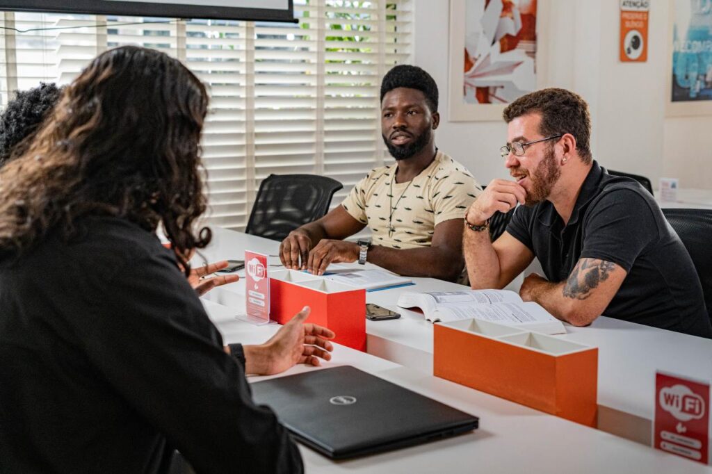 Imagem que representa a diversidade cultural nas empresas. Há 4 pessoas na foto, todos são migrantes e ex-estudantes da Toti Diversidade. Todos são do sexo masculino. Há duas pessoas negras, uma indígena e uma branca. Os 4 estão em uma mesa conversando. Dois deles estão de costas para a foto e um deles aparece somente o cabelo na foto. 