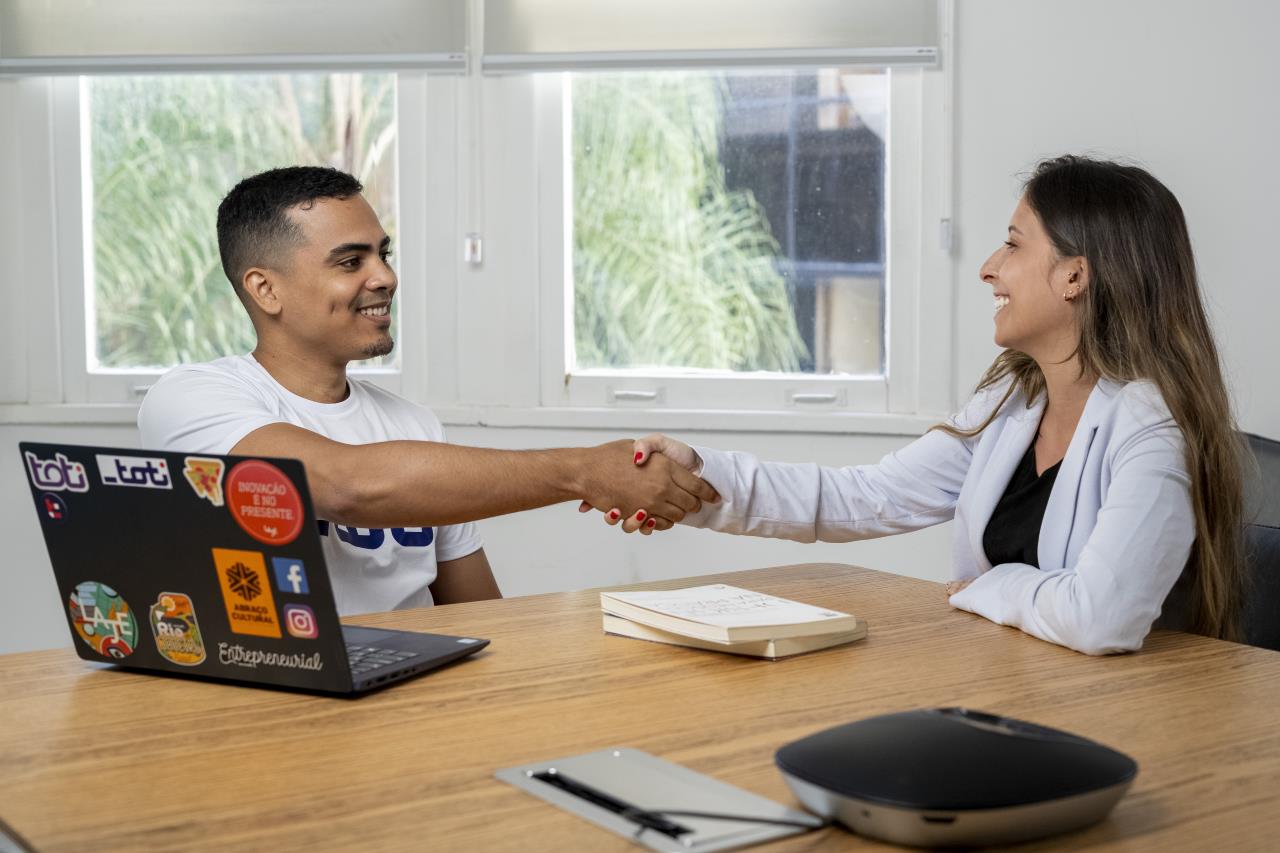 Imagem de um homem e uma mulher sorrindo e apertando as mãos sobre uma mesa de escritório.