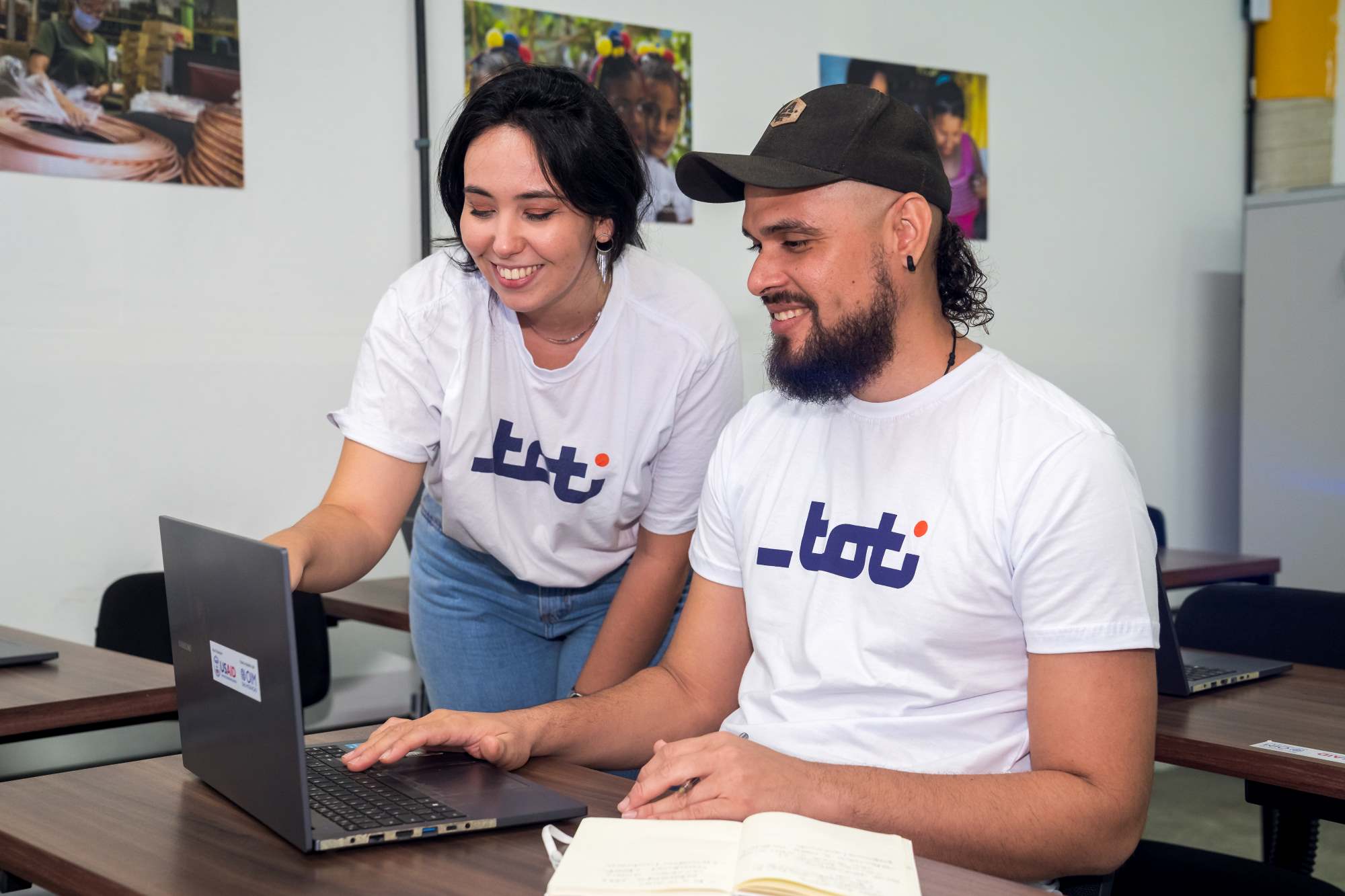 Foto de uma mulher e um homem sorrindo enquanto olham para um laptop, ambos vestindo camisetas brancas com o logo da Toti. Eles estão em um ambiente de sala de aula com fotos coloridas nas paredes.