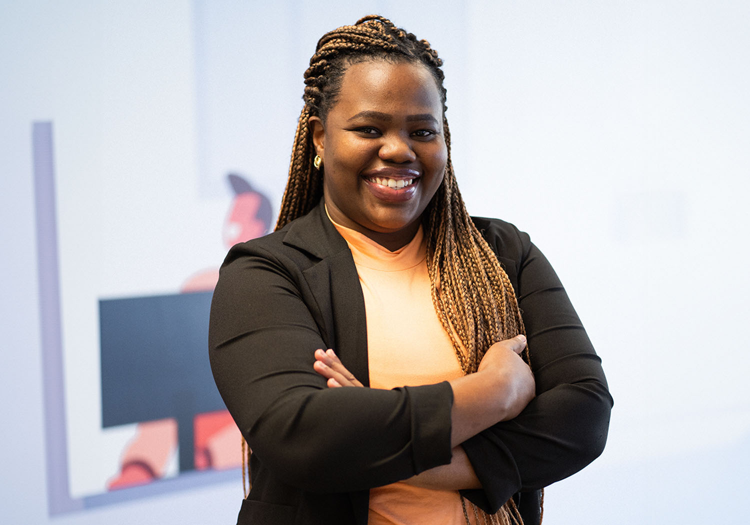 Mulher de braços cruzados sorrindo. Ela veste uma camisa laranja e um blazer preto formal.