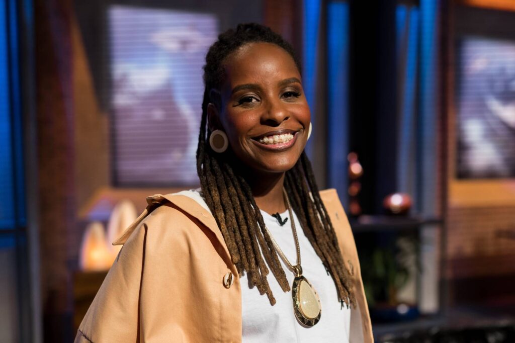 Monique Evelle, empresária, investidora e comunicadora, é uma mulher negra e está vestindo uma blusa branca e um casaco marrom. Está sorrindo.