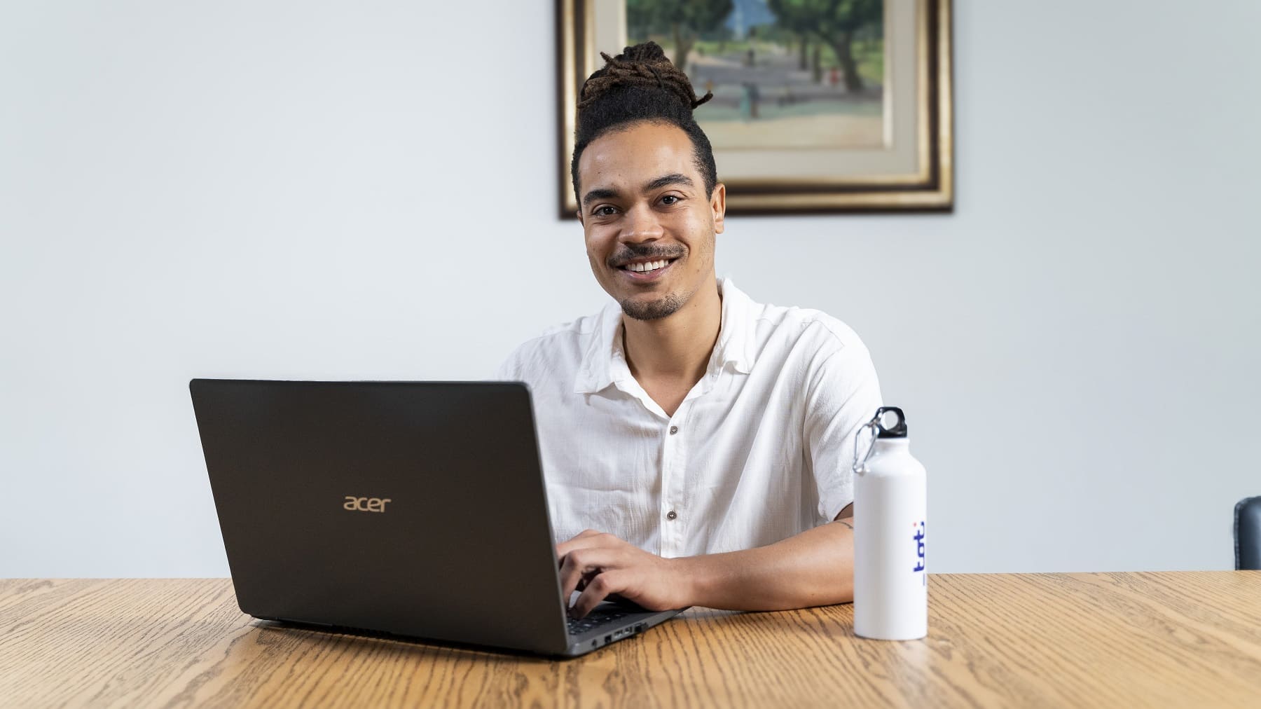 Um jovem homem negro está sentado utilizando um notebook. Em sua mesa, há uma garrafa branca. Ele sorri para a foto.