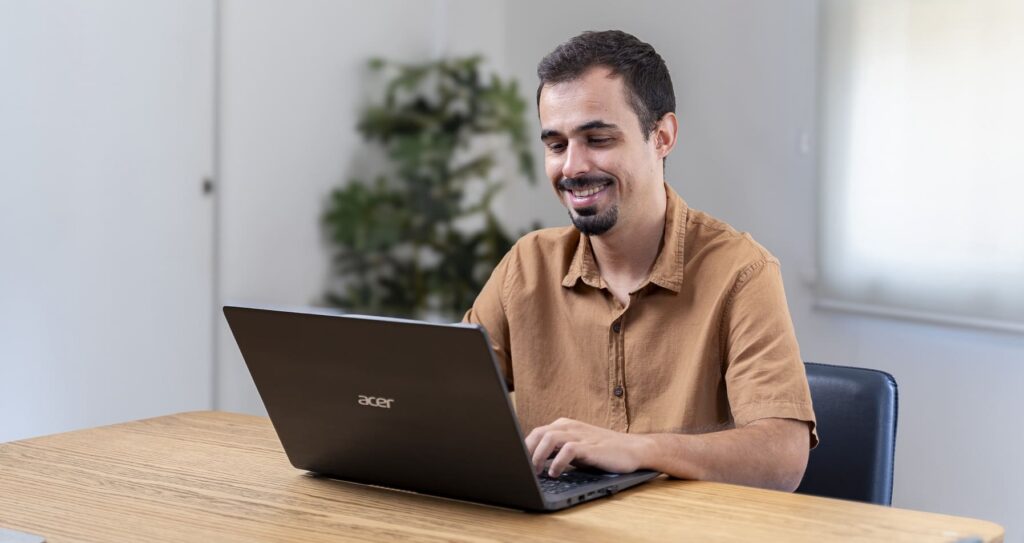 Pessoa do sexo masculino usando um notebook, o qual está apoiado em uma mesa. Essa pessoa possui cabelo preto, barba e veste uma camisa de manga. A imagem representa o trabalho remoto, que é uma das tendências de RH para 2024