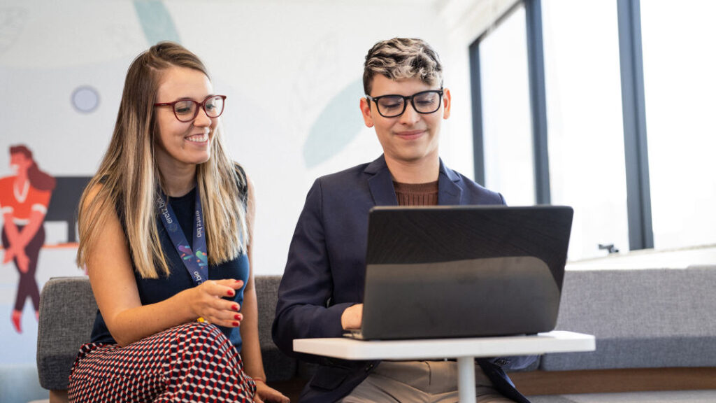 Duas pessoas estão sentadas. Uma delas está com um notebook e mostra à outra a sua tela. Ambos parecem estar em um ambiente corporativo.