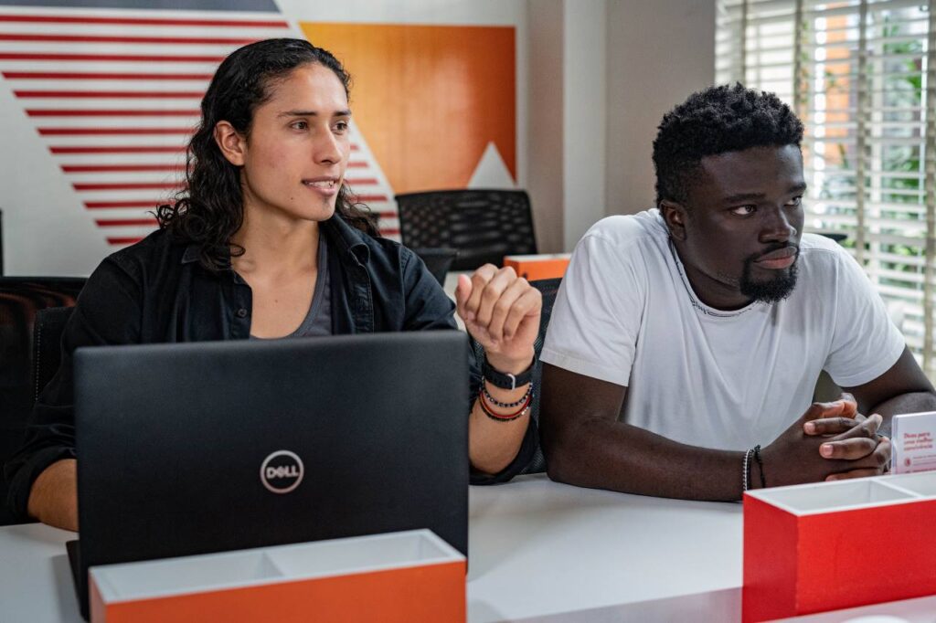 Há duas pessoas na foto: Bryan e Seth, ambos profissionais formados pela Toti Diversidade. Bryan é indígena e colombiano. Seth é negro e ganense. Ambos estão sentados em uma mesa. Bryan está usando um notebook.