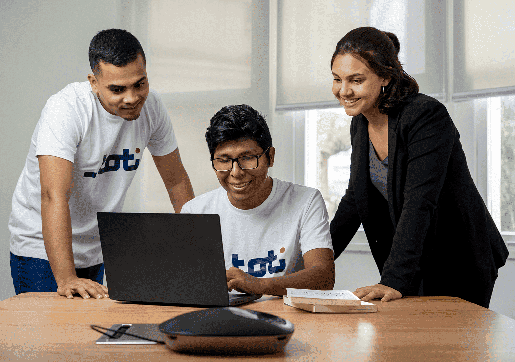 Um homem está sentado usando seu notebook, com um homem e uma mulher ao lado, auxiliando-o. Todos estão sorrindo. A imagem remete a uma ação de voluntariado empresarial.