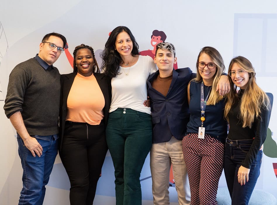 Seis pessoas estão posando para a foto. Todos estão abraçados e sorrindo. Há quatro mulheres e dois homens. 