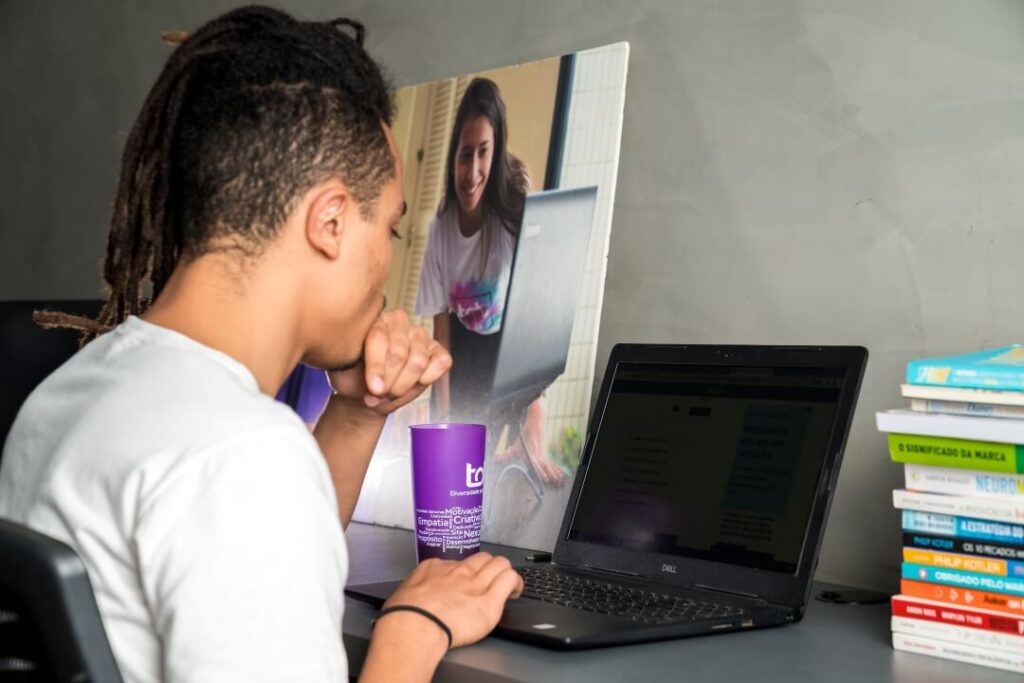 Jovem estudante com dreadlocks focado em seu laptop ao lado de uma pilha de livros, indicando dedicação ao seu trabalho.