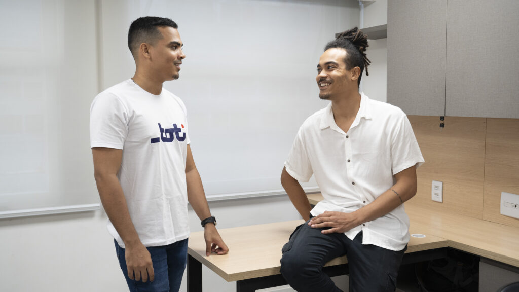 Exibe dois homens em um escritório, ambos sorrindo e em pé, um frente ao outro em uma conversa amigável. O ambiente é informal e amigável, refletindo um local de trabalho acolhedor e inclusivo.