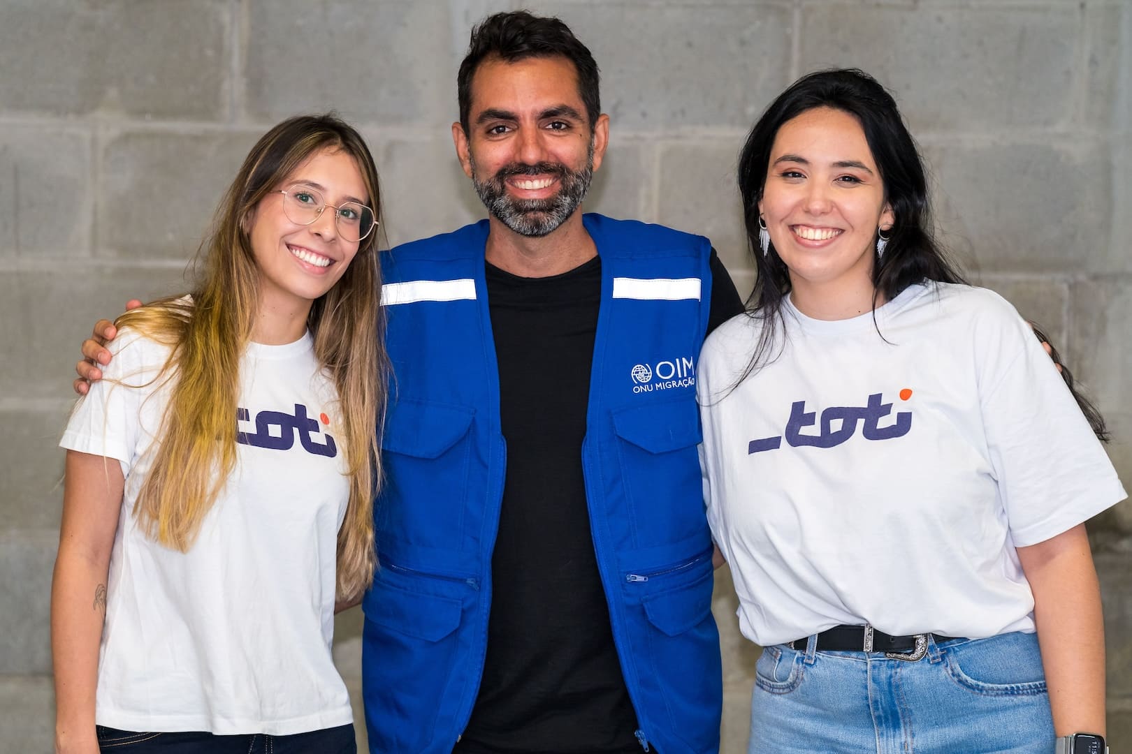 A imagem exibe três pessoas sorridentes. Duas mulheres, uma de cada lado, estão vestindo camisetas brancas com o logotipo da Toti, e um homem no centro usa um colete azul com o logotipo da OIM (Organização Internacional para as Migrações).