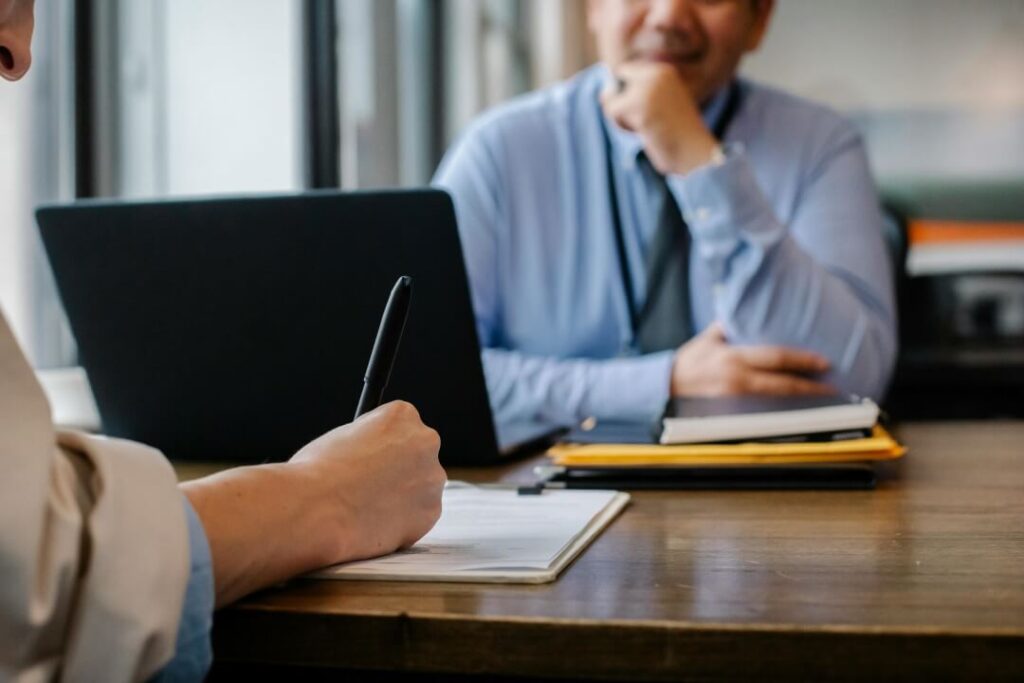 Em um escritório, uma candidata preenche um formulário de emprego sob a observação atenta de um entrevistador masculino ao fundo, enfatizando um momento crucial de uma entrevista de trabalho.