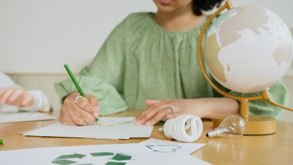 Mulher com uma blusa verde, escrevendo em um papel sobre uma mesa.