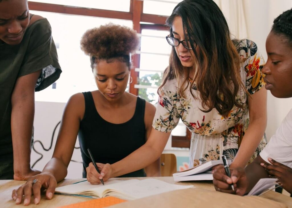 Duas mulheres trabalhando sobre uma mesa, enquanto outras duas pessoas observam.