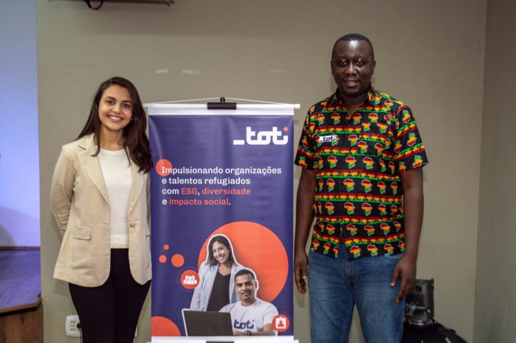Duas pessoas posando para foto com um banner da TOTI atrás.