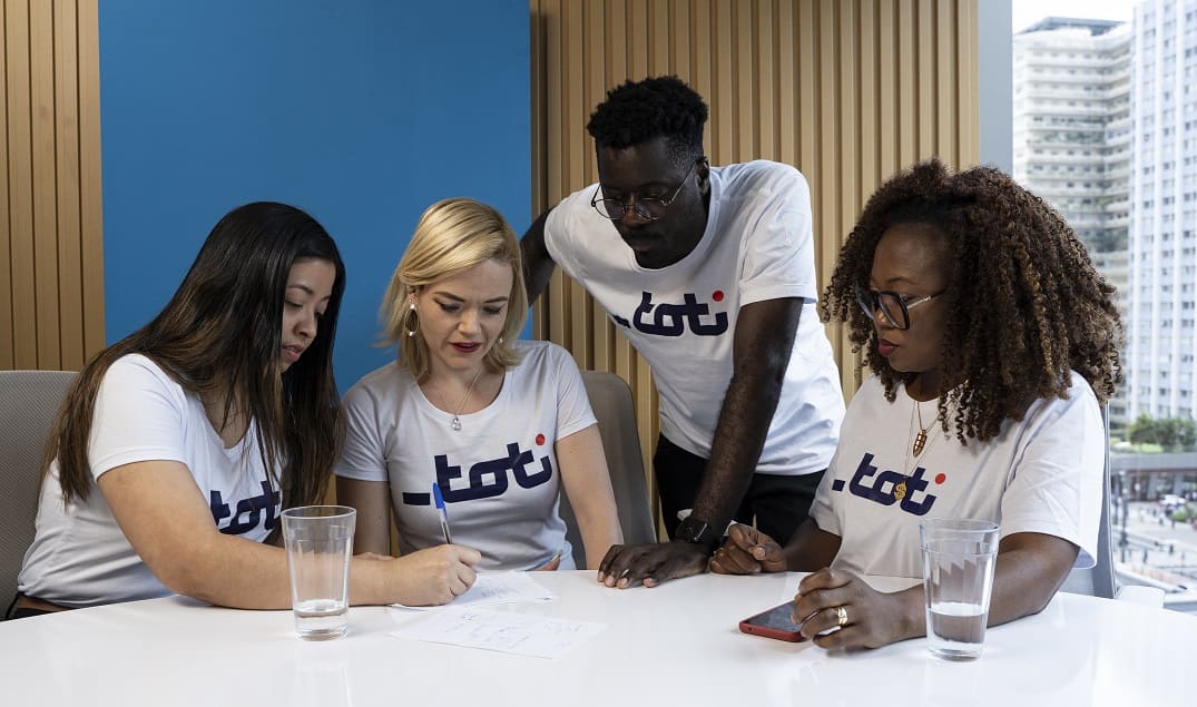 Grupo diverso de quatro pessoas usando camisetas da Toti Diversidade, reunidos em uma mesa de escritório e discutindo documentos, representando colaboração e inclusão no ambiente corporativo.