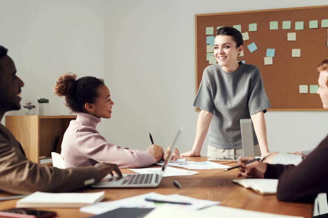 Sala com mulheres sentadas a mesa e uma em pé dialogando.