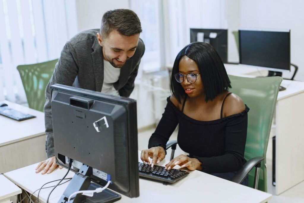 Uma mulher sentada a mesa em frente ao computador com um homem ao lado auxiliando.