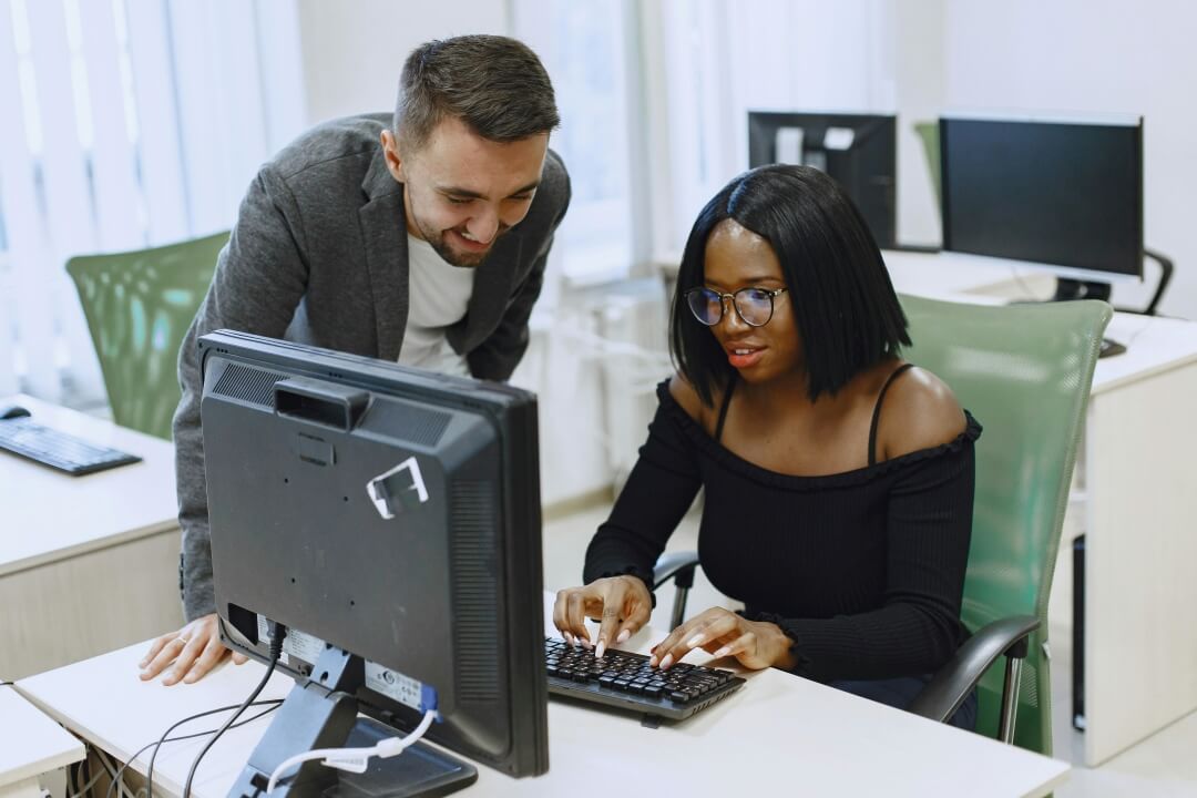 Uma mulher sentada a mesa em frente ao computador com um homem ao lado auxiliando.