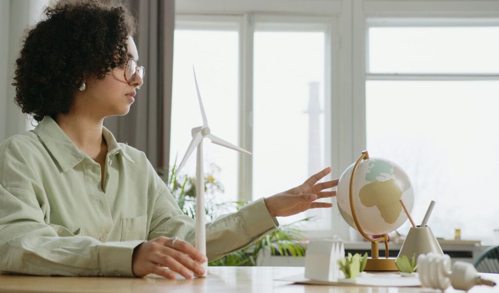 Pessoa sentada à mesa em um ambiente iluminado, observando um globo terrestre enquanto segura um modelo em miniatura de turbina eólica. Sobre a mesa há objetos relacionados à sustentabilidade, como pequenas plantas e uma lâmpada, simbolizando práticas de ESG.
