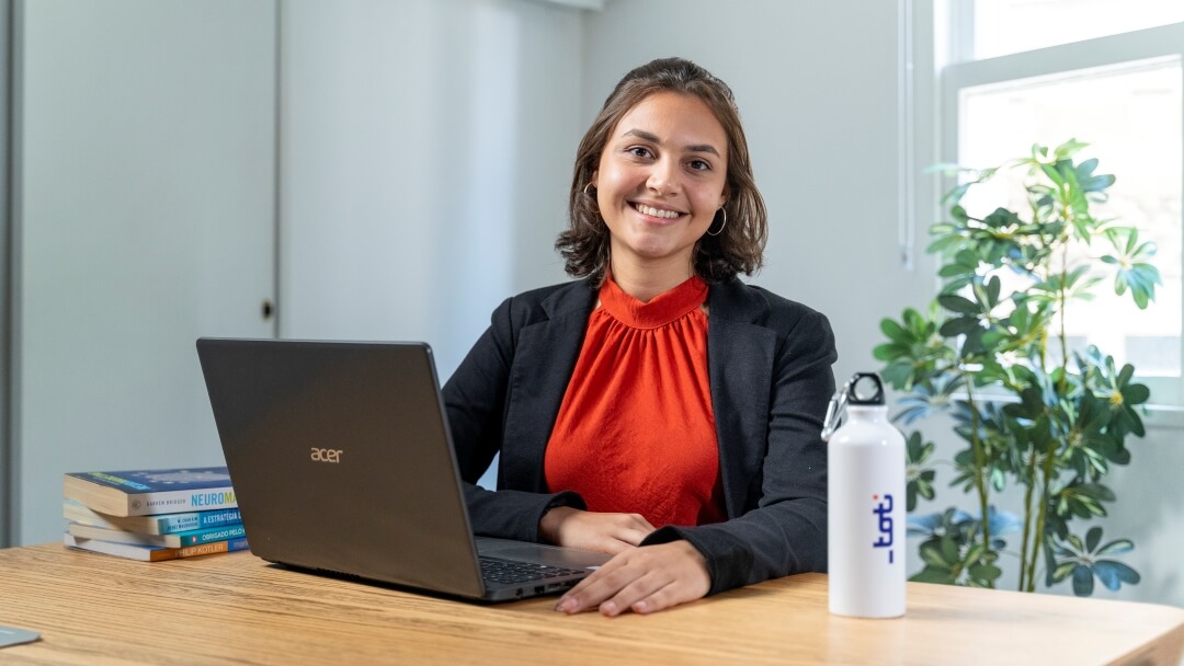 Mulher sentada a mesa posando para a foto com um notebook a sua frente.