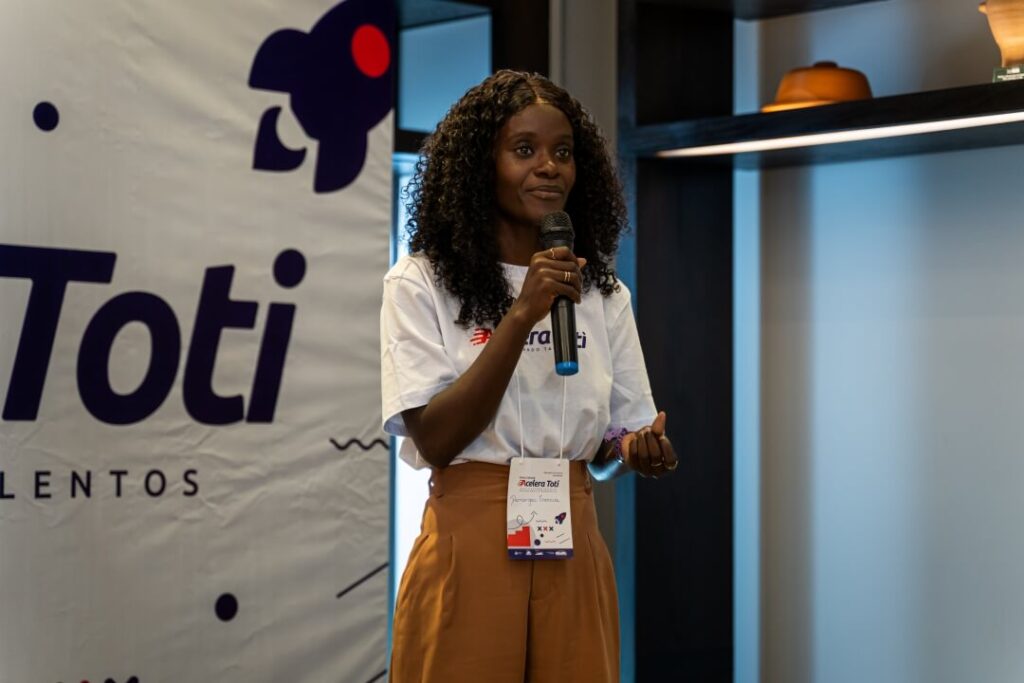 Mulher em pé, palestrando, com imagem da logo da Toti ao fundo.