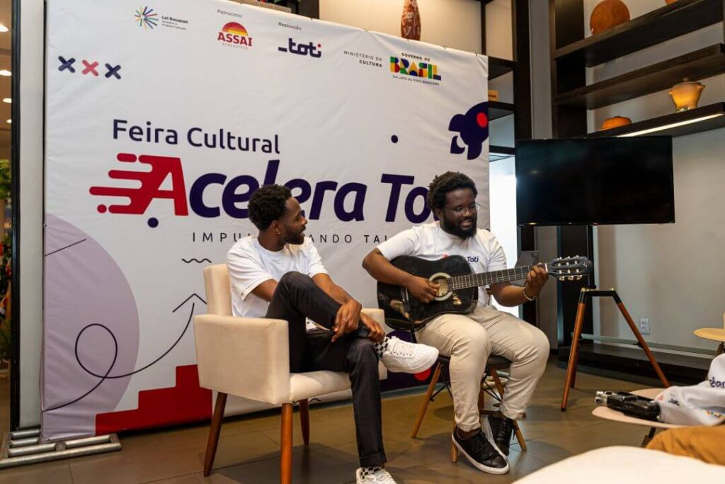 Duas pessoas sentadas em uma apresentação, uma delas está tocando violão.