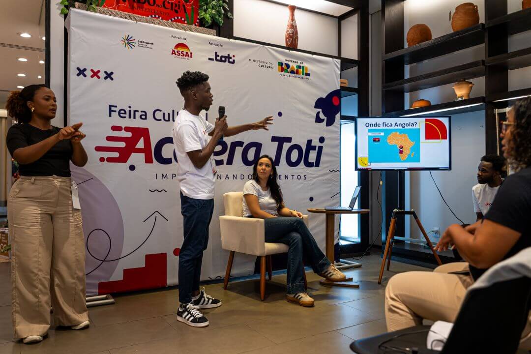 Duas pessoas realizando uma palestra, uma esta em pé e a outra sentada.