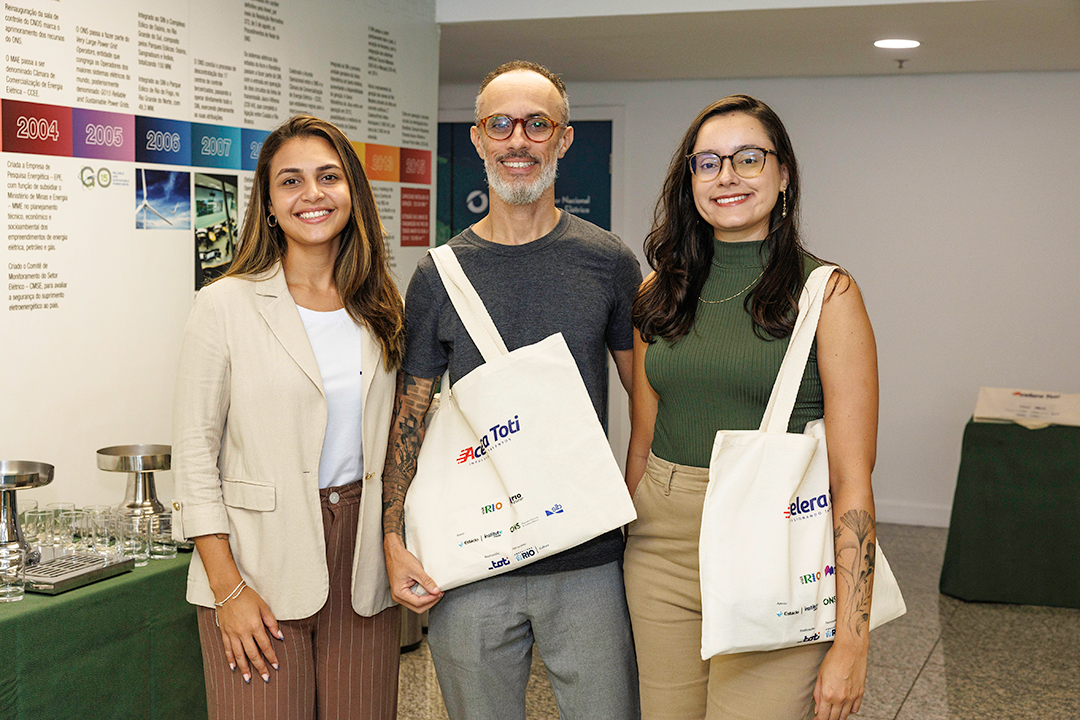 Três pessoas sorrindo posam em ambiente interno; duas delas seguram ecobags brancas com logotipos, durante um evento ou encontro corporativo.