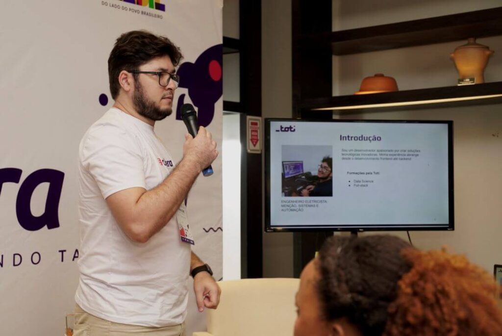 Homem em pé ao lado de uma televisão fazendo uma apresentação ao microfone.